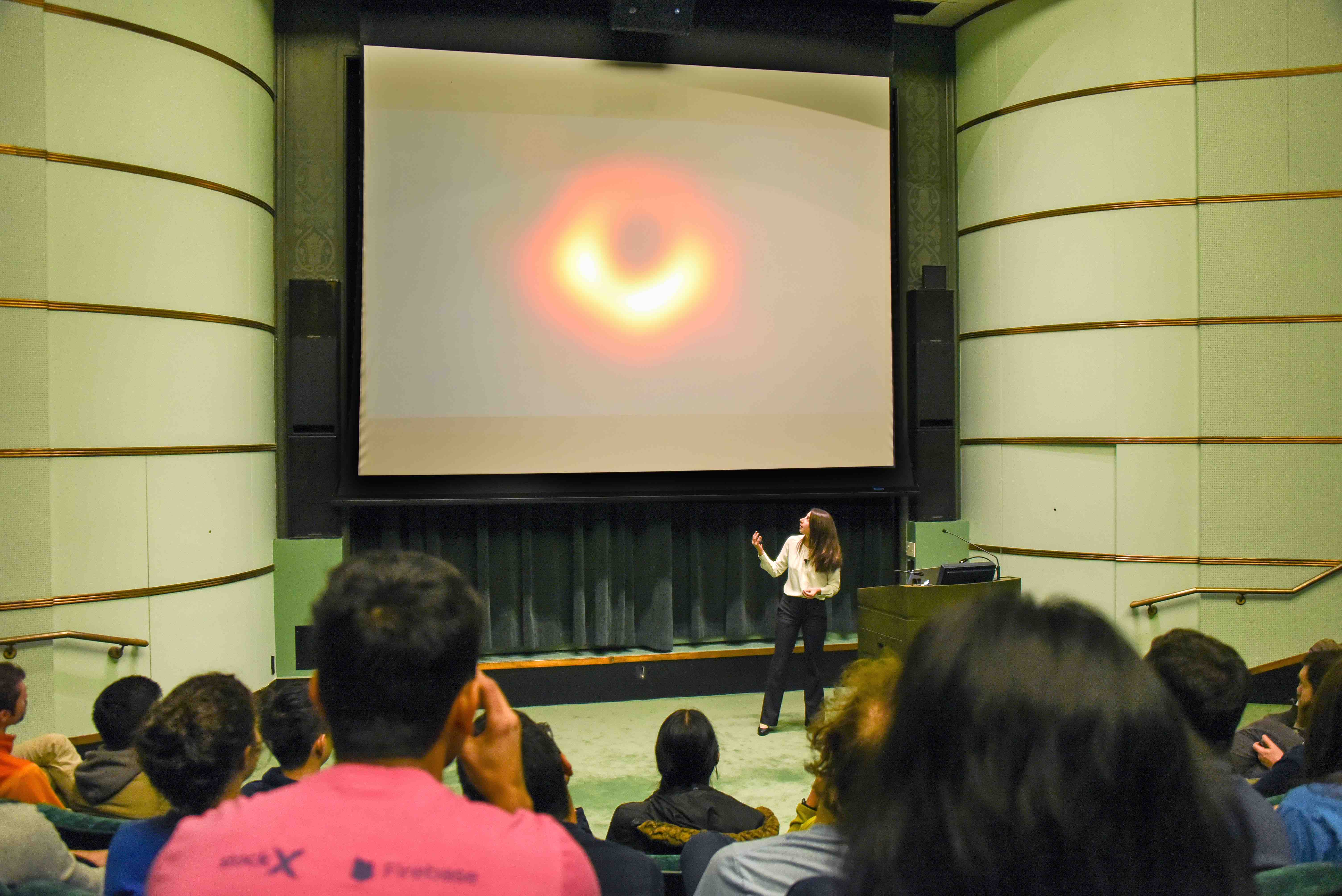 Dr. Bouman shows the first ever image of a black hole - a red/orange ring that's brighter on the bottom than the top