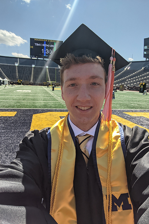 Matthew Lamb in grad cap, gown, and sash on the field of the Big House