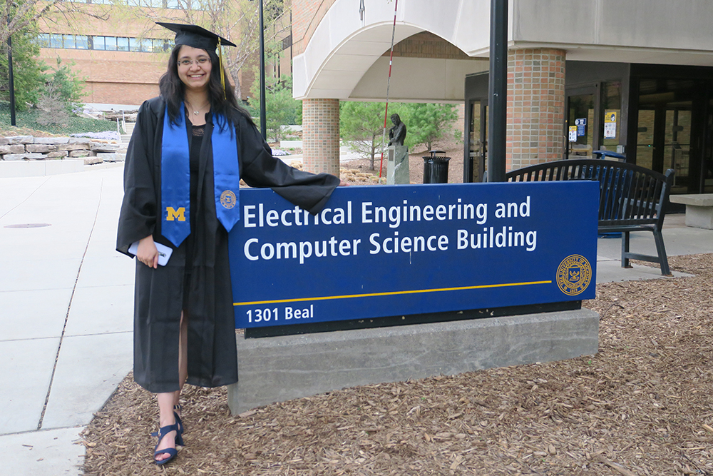 Rucha stands in grad cap and gown next to the EECS building sign