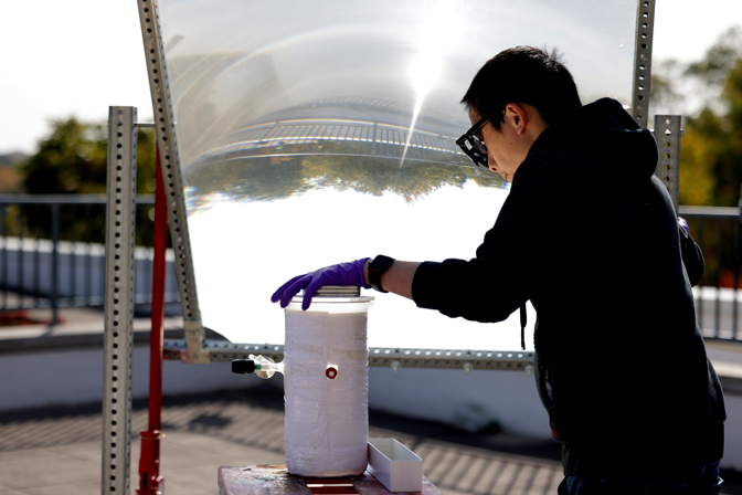 A researcher stands outdoors with the device to boost efficiency. It's a sunny day and the researcher is wearing safety gloves and glasses