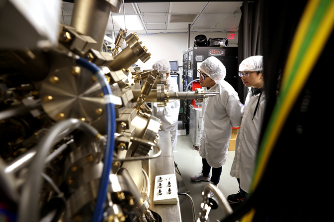 Researchers standing in lab coats and lab safety gear stand and observe a large machine growing nanowires.