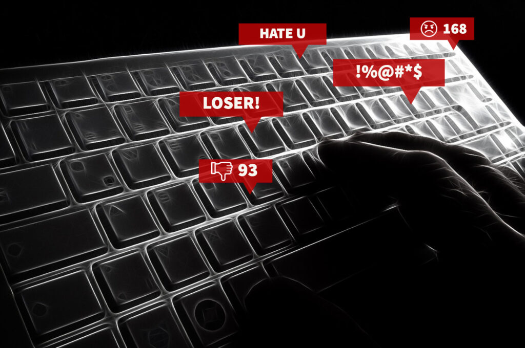 Black and white closeup of a hand on a computer keyboard surrounded by several red speech bubbles with examples of hateful speech (a thumbs down, an angry face, special characters implying cursing, etc.)