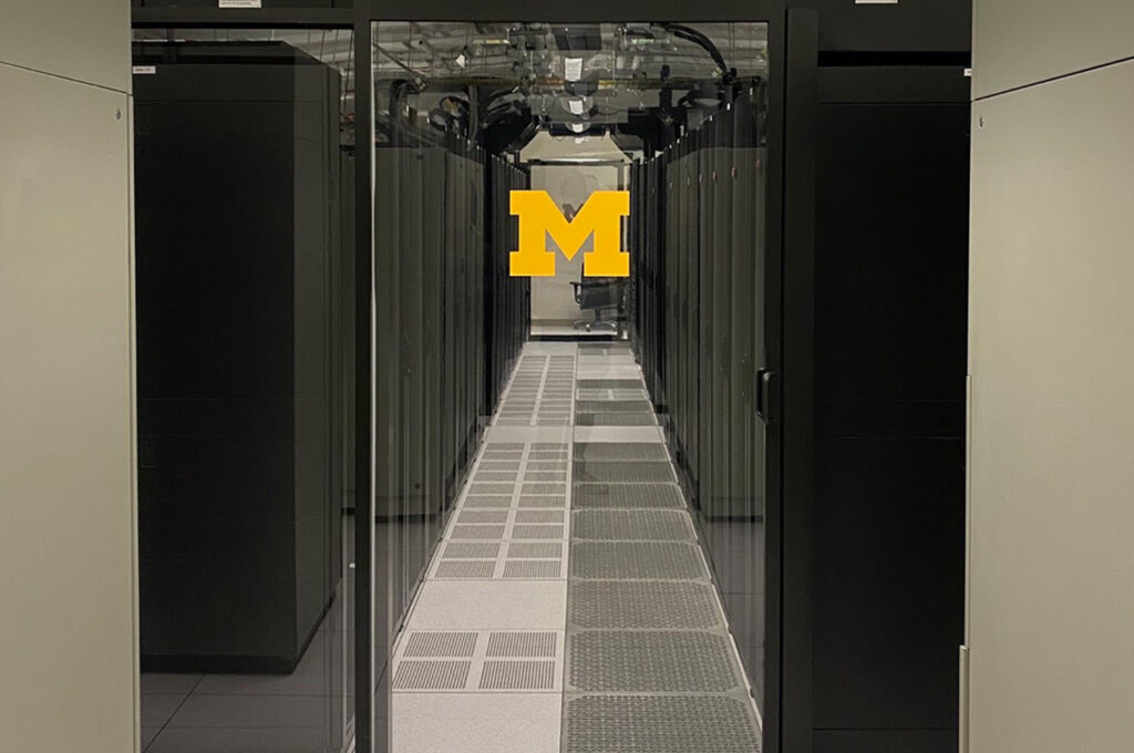 A glass door emblazoned with the U-M block M leads to a server room with black computer towers arranged in neat rows.