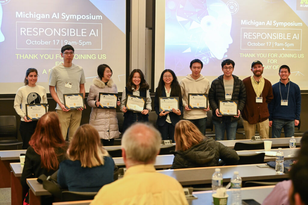 The award winners standing in a row in front of a lecture hall, holding their award certificates and smiling.