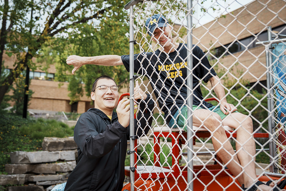 A student celebrates next to the dunk tank holding the ball that successfully dunked Prof. Fessler. Fessler smiles but gives a thumbs down behind the student.