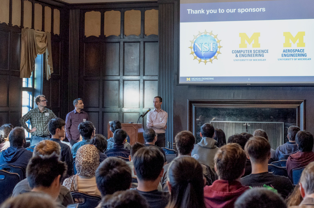 Prof. Jean-Baptist Jeannin speaks at a lectern in a large wood-paneled room before a large group of people seated in chairs. Event organizers Cyrus Omar and Max New stand to his side. Behind him is a screen with a powerpoint slide thanking the event sponsors, the NSF, U-M CSE, and U-M Aerospace.
