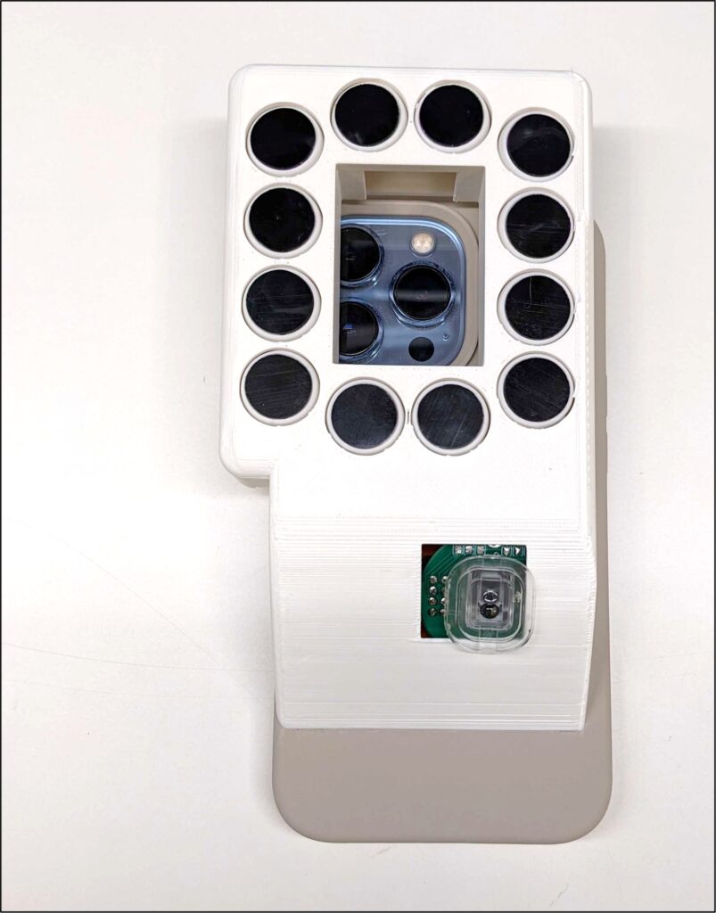 The picture shows the underside of BrushLens, a white phone case. A window in the center of the case reveals a smartphone camera. Thirteen black circles, which are the clickers, surround the window in a circle.