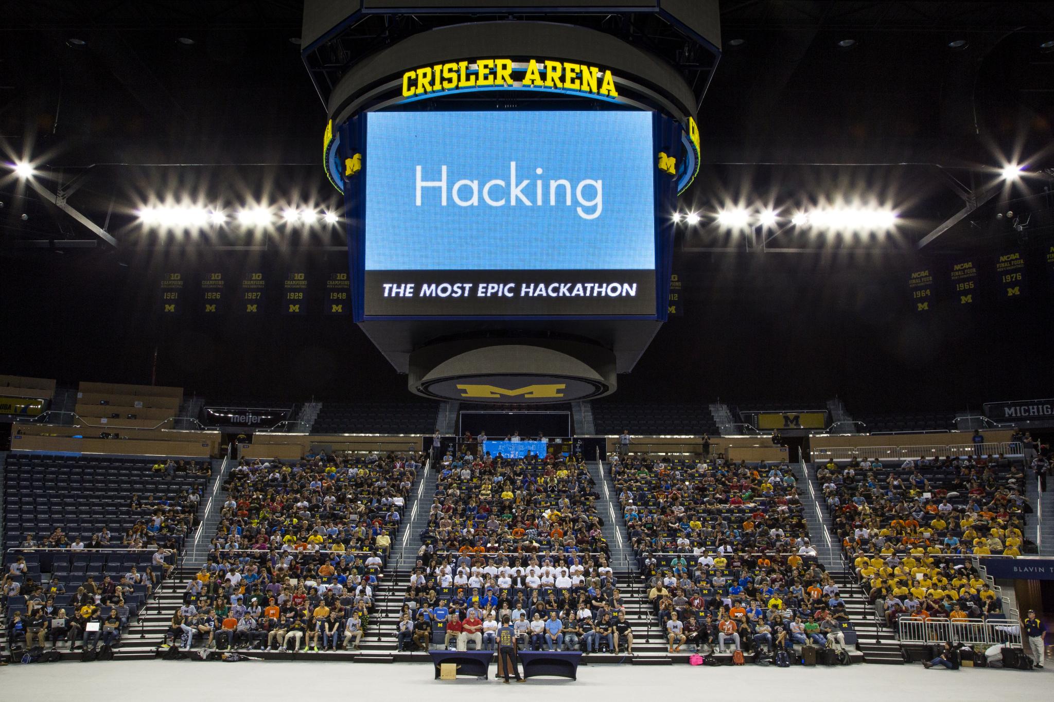 A person stands at a table and speaks to a crowd of students at the MHacks opening ceremony.