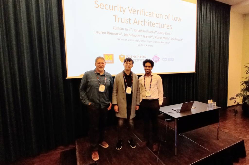 CSE Prof. Todd Austin, Princeton PhD student Qinhan Tan, and CSE PhD student Yonathan Fisseha stand in front of screen with a black curtain behind it. The title of their paper and school logos appear on the screen. 