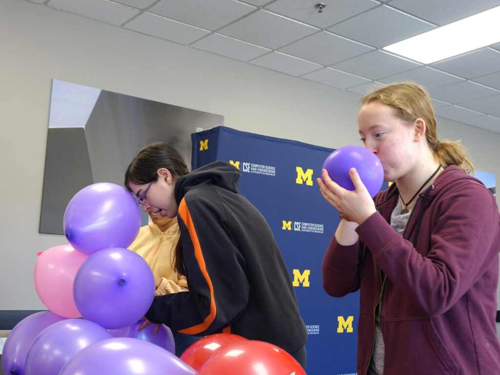 Two students blow up pink and purple balloons lined up on a table.