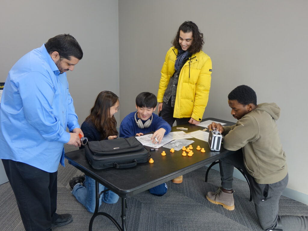 Five students kneel and stand around a table. Two of them are working together to fill out a worksheet. There is a collection of about a dozen rubber ducks on the table.