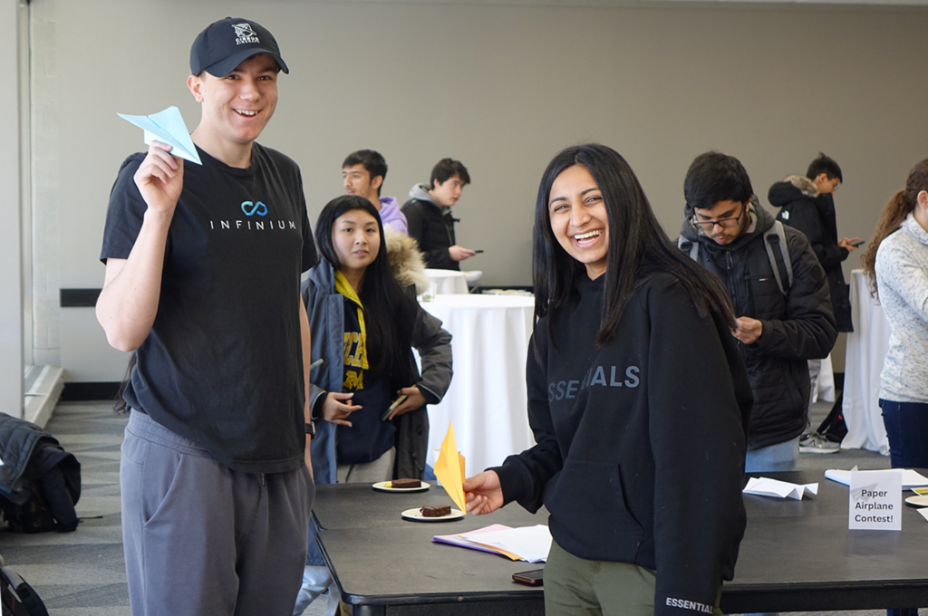 Two students, smiling, hold paper airplanes. Other student are milling and mingling in the background.
