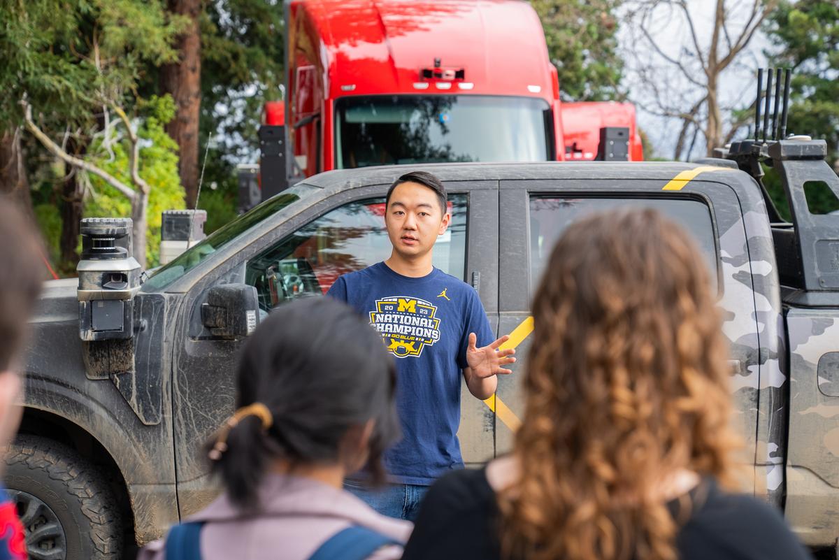 Students listen to a presenter in front of a black camo pickup truck and a red semi truck.