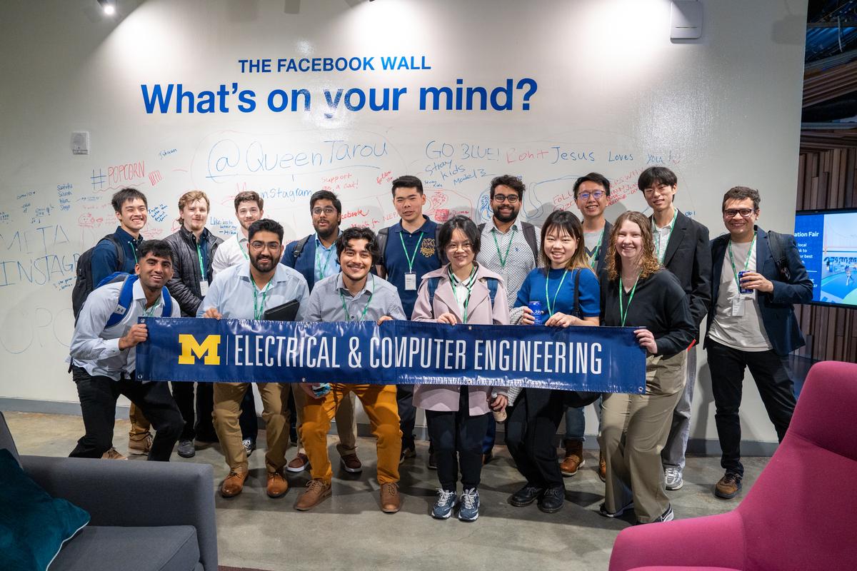 Students pose with the University of Michigan Electrical & Computer Engineering banner in front of a whiteboard wall labeled "The Facebook Wall" with the words "What's on your mind?"