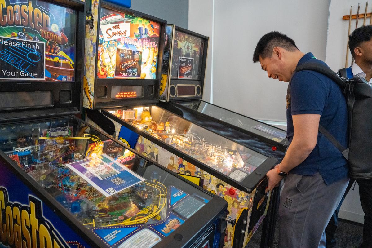 One student laughs while playing a "The Simpsons" pinball game. Two other pinball games are on each side of the student.