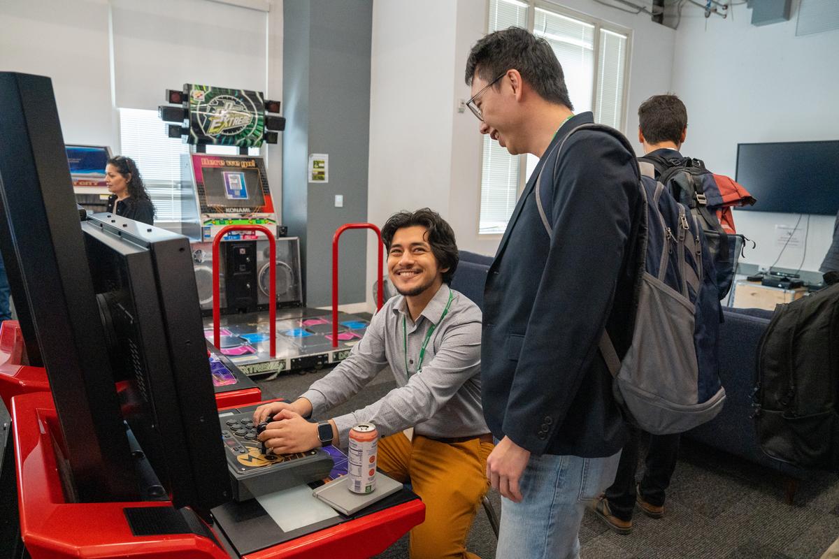 Two students smile as one watches the other play an arcade game on the Meta campus.