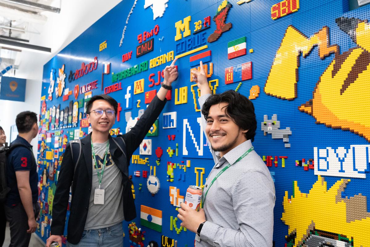 Two students stand against a blue background lego wall, filled with colorful logos from universities and companies. The students point at the Block M and the words "GO BLUE", above their heads.