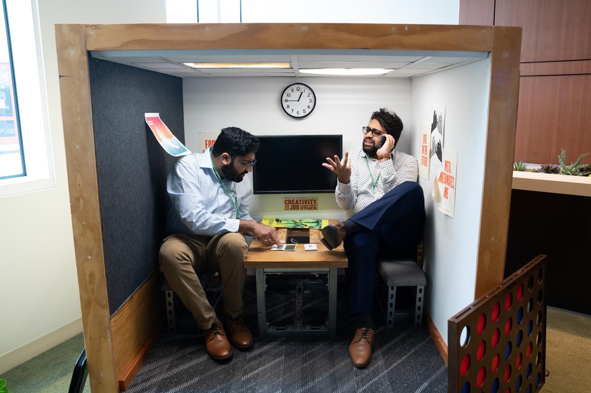 Two students pretend to work in a miniature replica of a Meta office, complete with a clock, posters, a tiny table, and TV screen. One student pretends to use a palm-sized laptop while the other has an animated phone call.