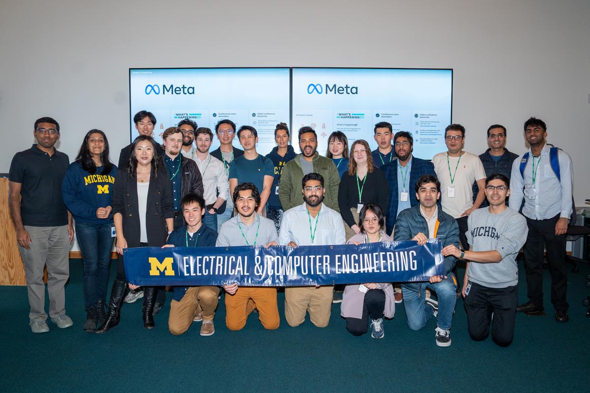 A group of students and Meta engineers pose for a photo with the Michigan Electrical & Computer Engineering Banner in front of two screens with the Meta logo.
