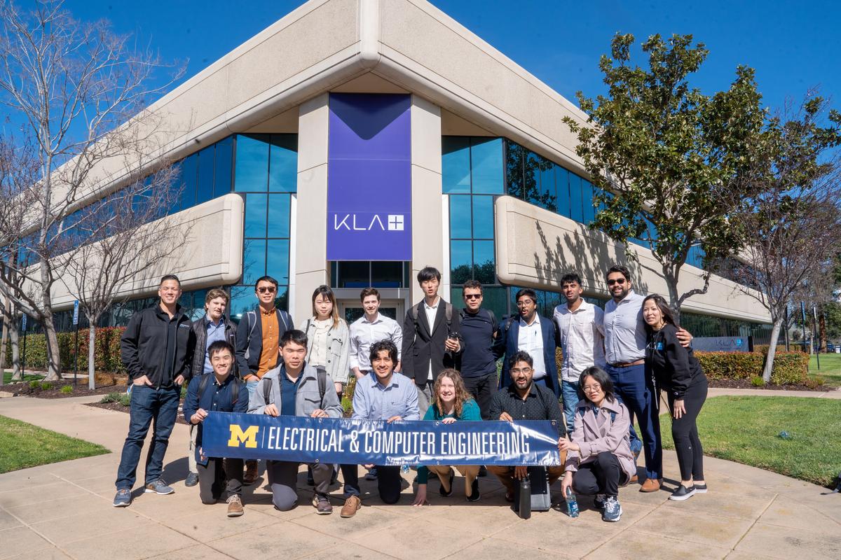 Students and KLA engineers pose in front of the KLA building, a triangular concrete corner with blue reflective windows. They are holding the Michigan Electrical & Computer Engineering banner.