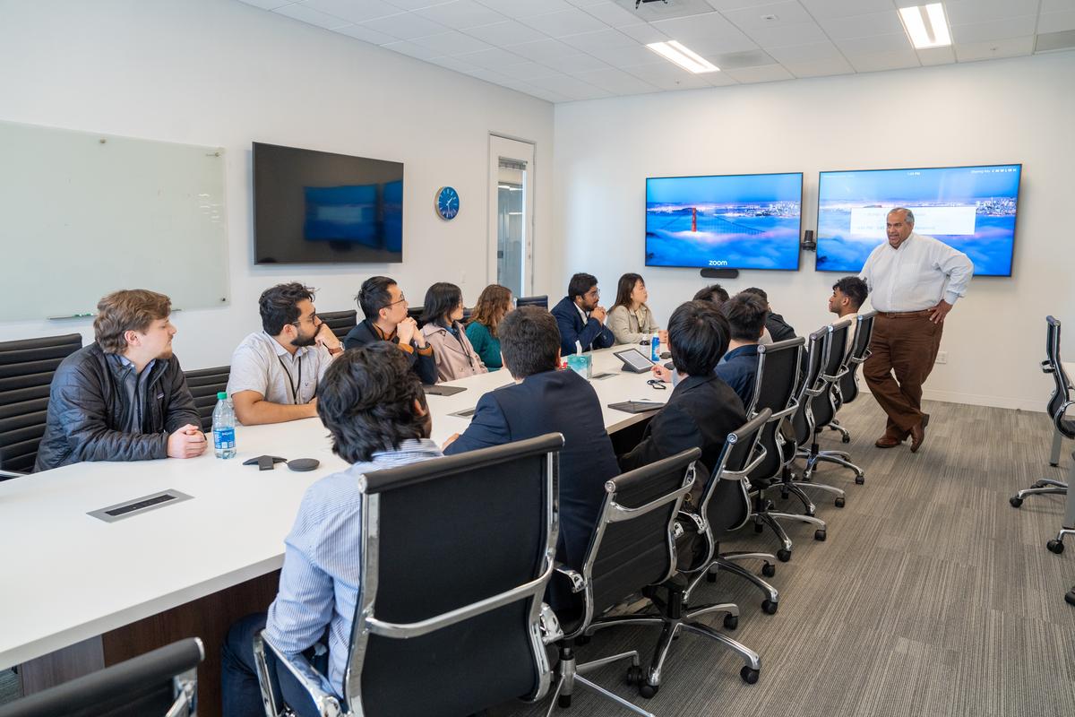 Students sit around a long table in rolling office chairs, listening to a presentation by alum Piyush Sevalia, who is in front of two large TV screens.