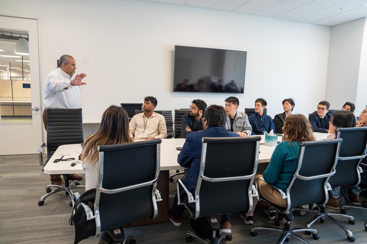 Students sit around a long table in rolling office chairs, listening to a presentation by alum Piyush Sevalia, who is gesturing during an explanation.