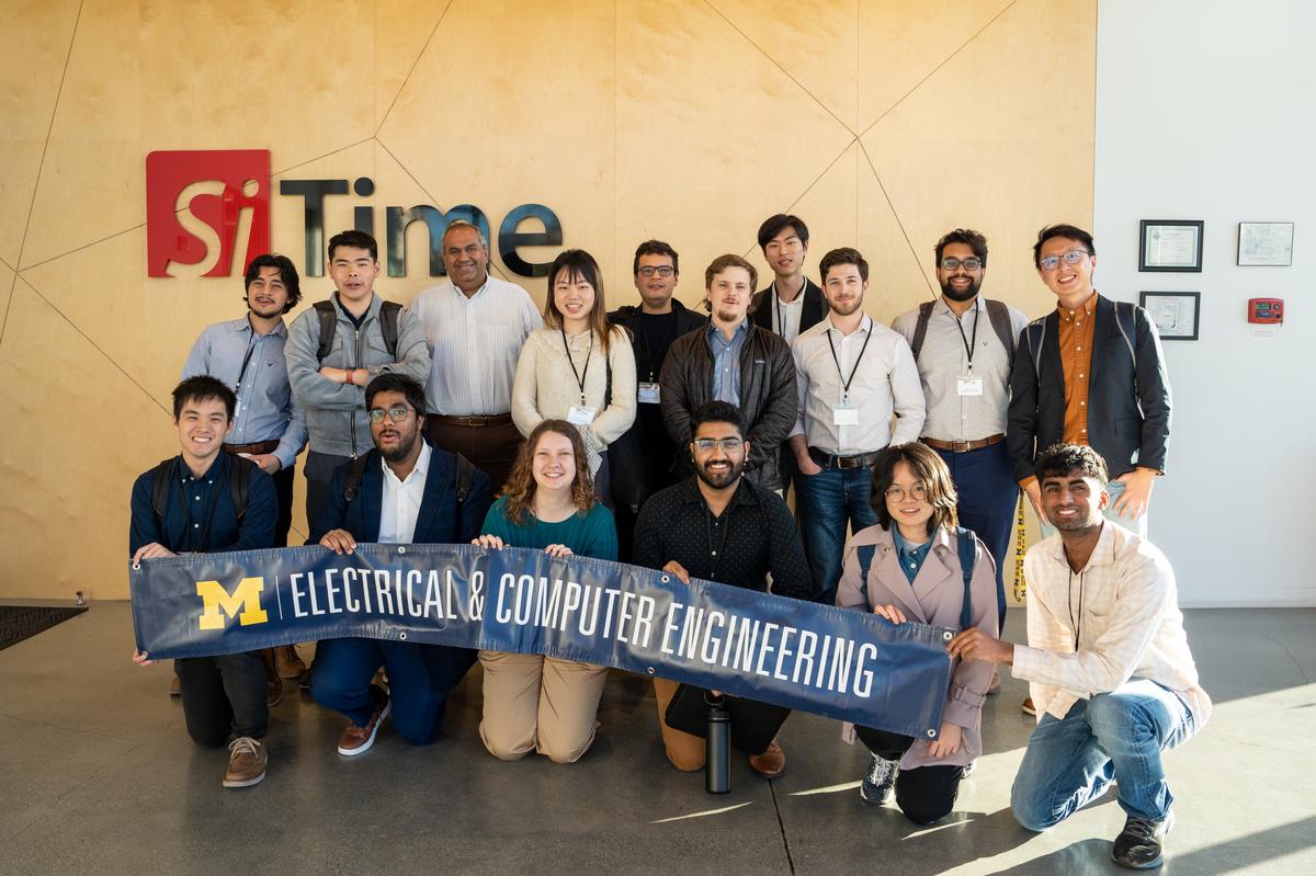 A group of students poses with alum Piyush Sevalia in front of a wall with the SiTime logo - a red box with Si in negative space, next to the black word Time.