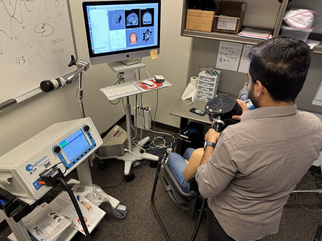 A view from the back of Hammad Ahmad performing TMS on Priscila Santiesteban. Ahmad is holding a black device about the size of a hairdryer up to Santiesteban's head. The device is hooked up to a box-shaped machine that is generating an electric pulse. Ahmad looks at a computer screen where a scan of Santiesteban's brain is projected.