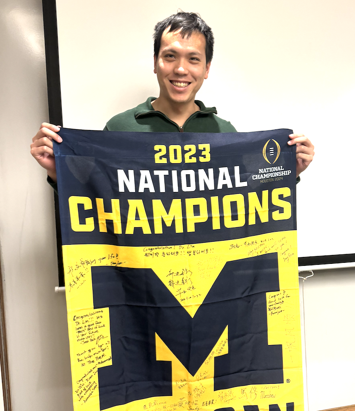 Wei-Kuan Lin, a Taiwanese man with short, black hair, stands wearing a green quarter-zip jacket. He is smiling and holding up a University of Michigan flag that says 2023 National Champions and has been signed by his friends and colleagues.
