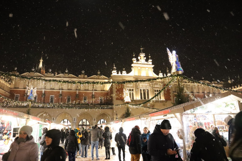 An ornate building is adorned with garland and Christmas lights. In front of the building are lit stalls selling Christmas-related treats and goods. Dozens of people are visiting the stalls. It's lightly snowing.