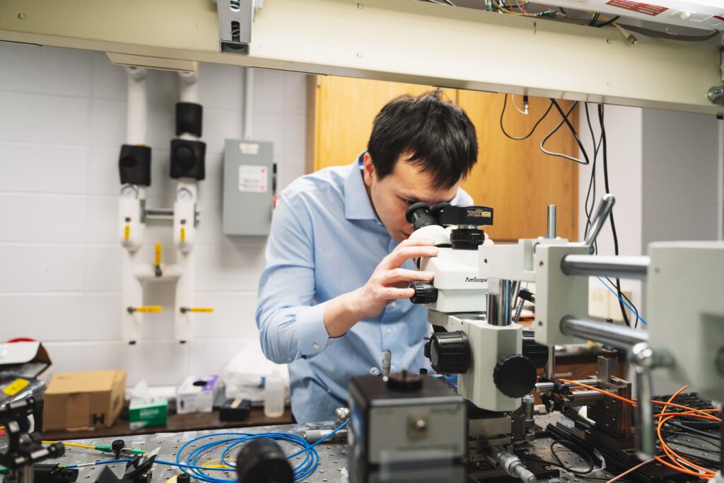 Wei-Kuan Lin, a Taiwanese man with short black hair, wearing a light blue button up shirt, looks into a microscope. He is in a laboratory setting, surrounded by electronic equipment.