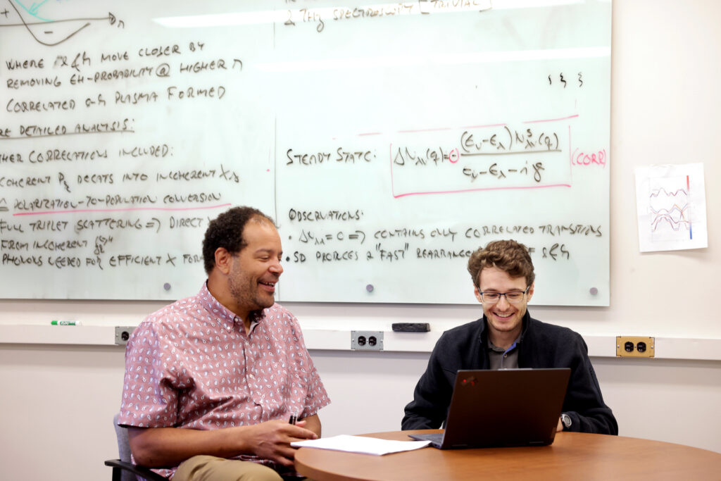 Markus Borsch and Mack Kira sit at a table, looking at a laptop and smiling. Behind them, a whiteboard is filled with physics equations and notes.