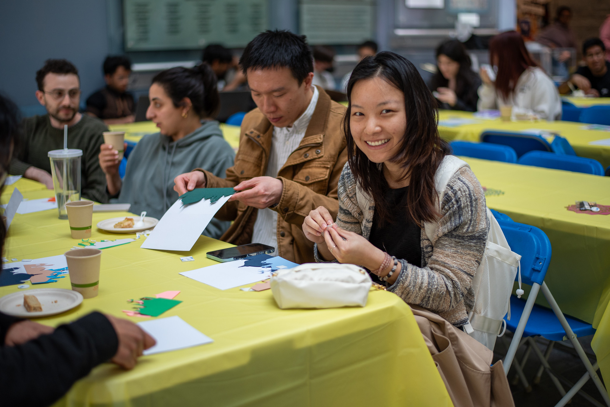 Students sit at a long table with a yellow tablecloth. They are working on craft, attaching colorful paper cutouts to cards. The student on the right is smiling  at the camera.