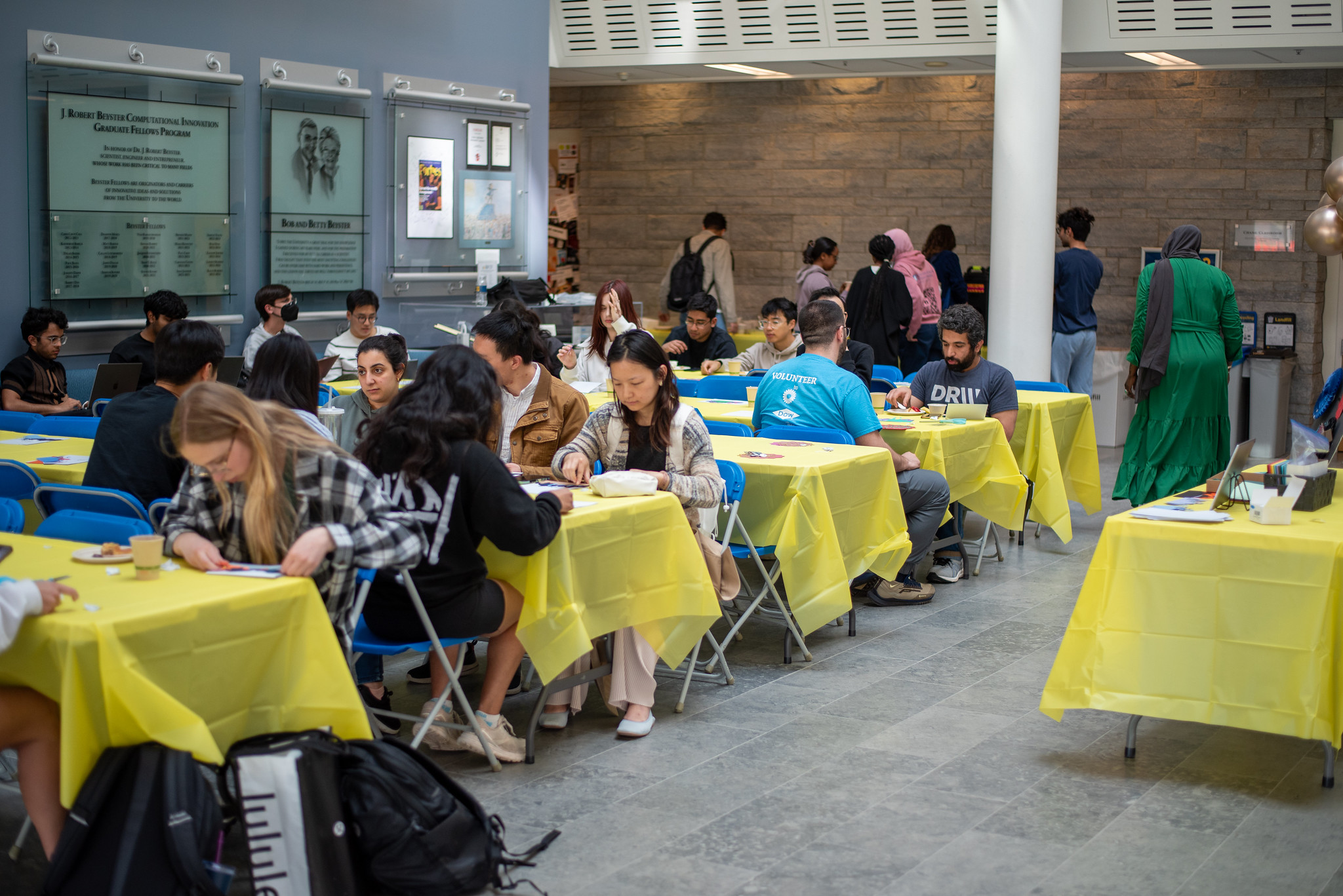 Several long tables with yellow tablecloths. About 20 people are seated at the tables working on crafts and eating snacks.