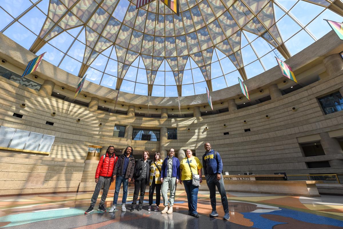 A diverse group of seven people stands in the center of the round entrance to the museum, with the arched glass ceiling above them.
