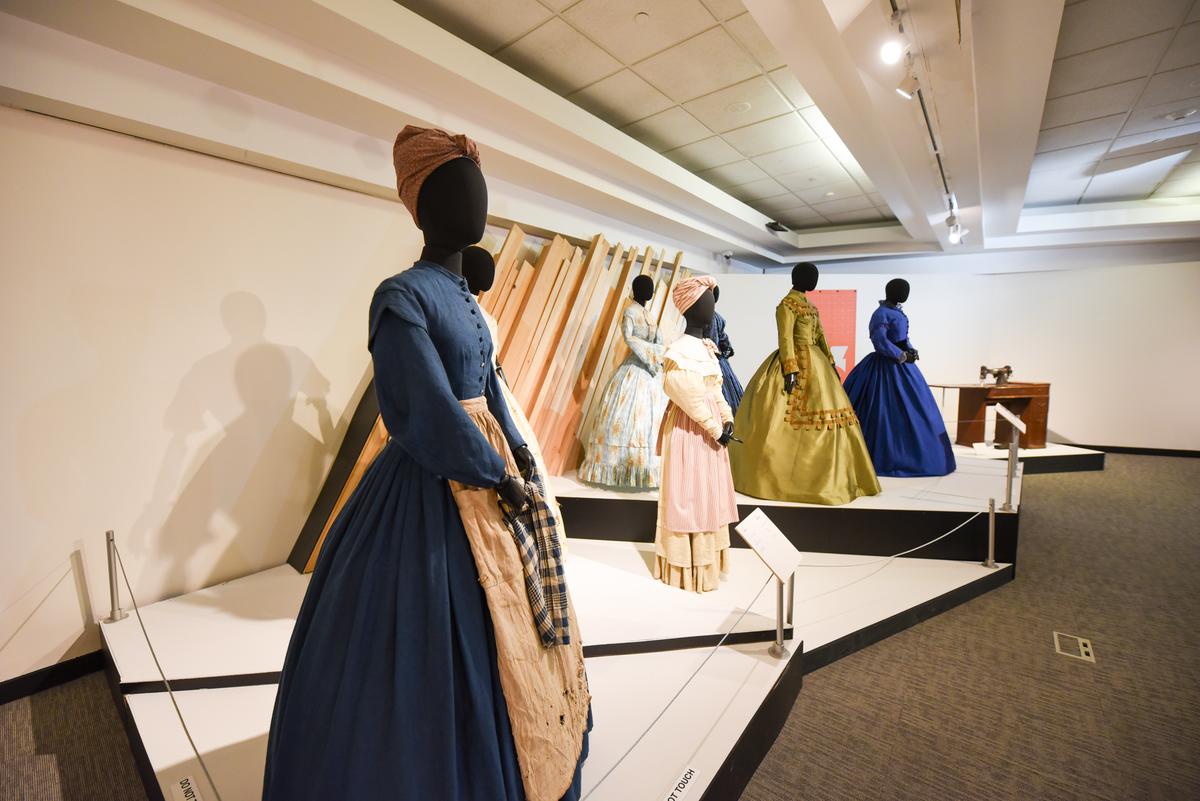 The exhibit by Ruth E. Carter, featuring costumes from the 2016 reboot of Roots. In the foreground, a mannequin with a navy blue long-sleeved dress and a beige apron, wearing a brown head wrap. Other costumes can be vaguely seen in the background of the photo, with similar clothing.