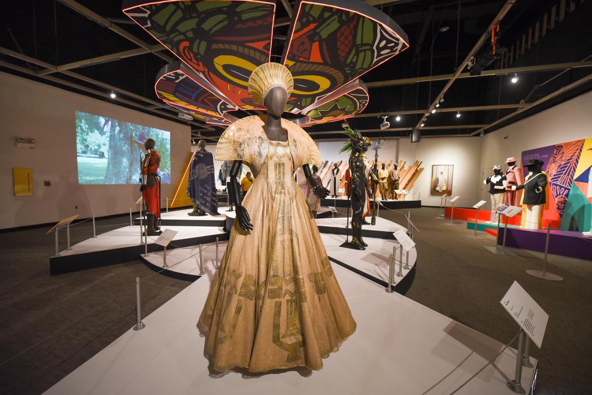 The exhibit by Ruth E. Carter, featuring costumes from the Black Panther movies. In the foreground, a mannequin with a matching beige dress and head piece. Other costumes can be vaguely seen in the background of the photo.