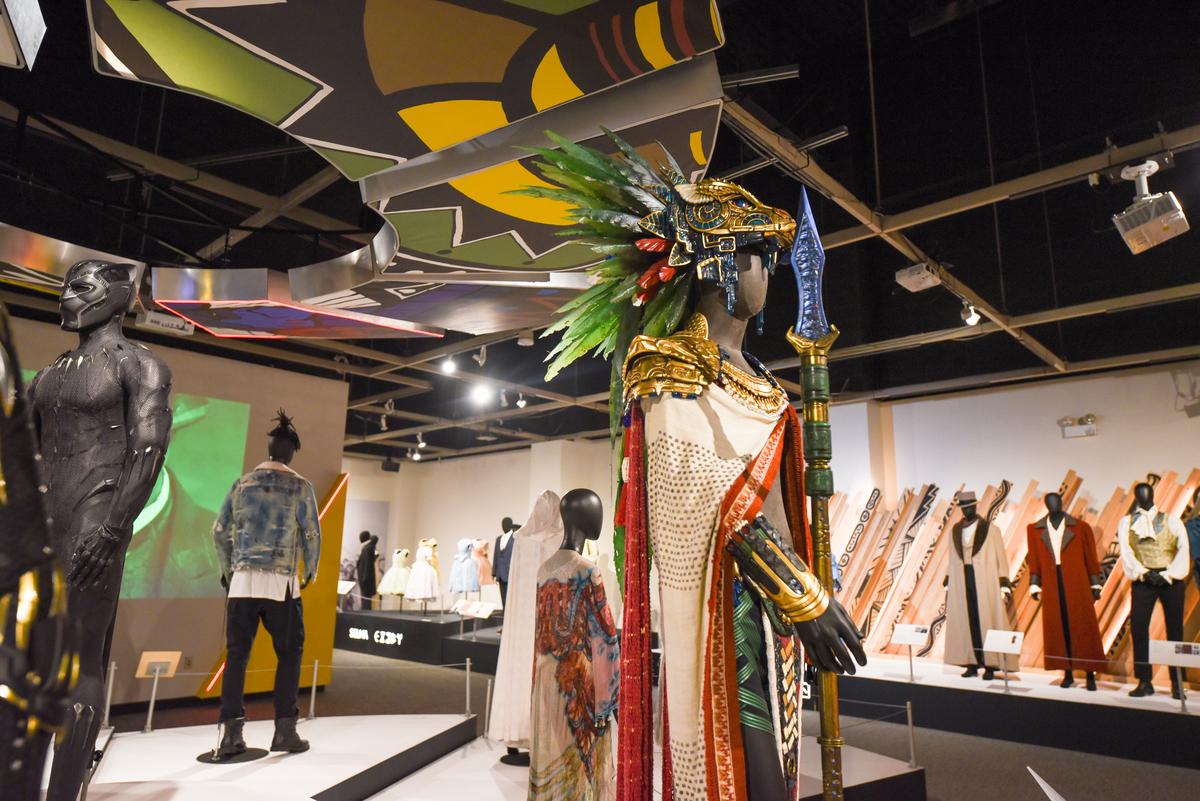 The exhibit by Ruth E. Carter, featuring costumes from the Black Panther movies. In the foreground, a mannequin with a white and red cloak, wearing a head piece shaped like a lion, with a mane of green and red feathers. The mannequin holds a spear. Other costumes can be vaguely seen in the background of the photo.