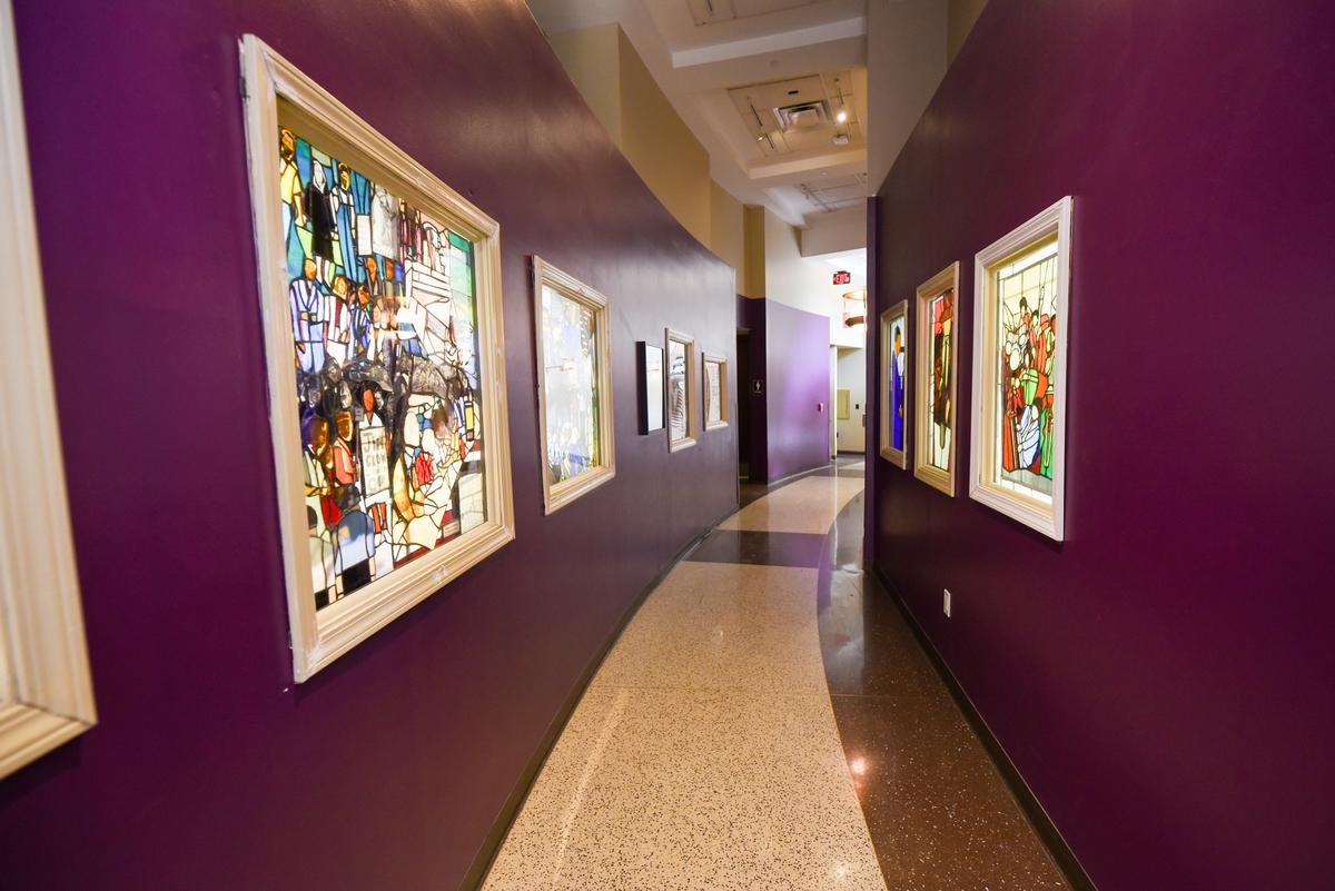 An eggplant-purple hallway is lined with colorful, stained glass windows on both sides. In the image, you can't quite make out what the figures in the glass are.