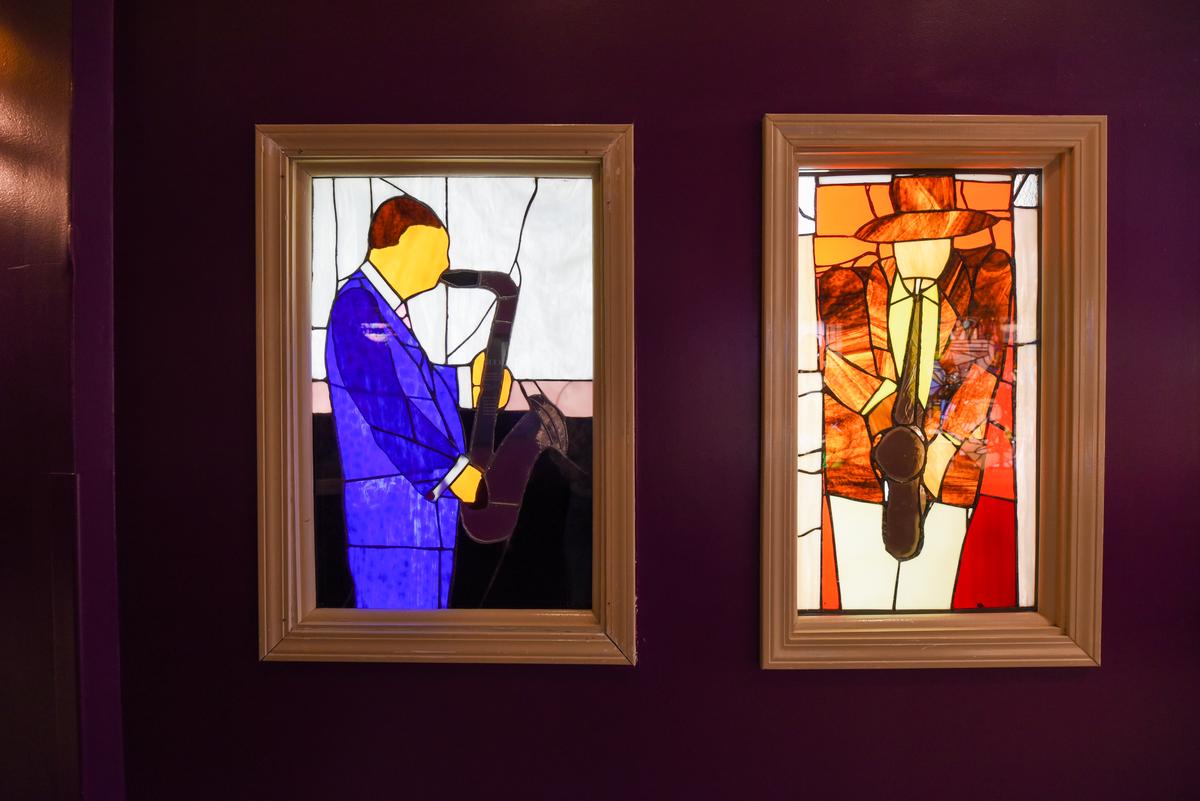 Two stained glass windows depict a person in a blue suit and a person in an orange coat and hat, both playing saxophones.