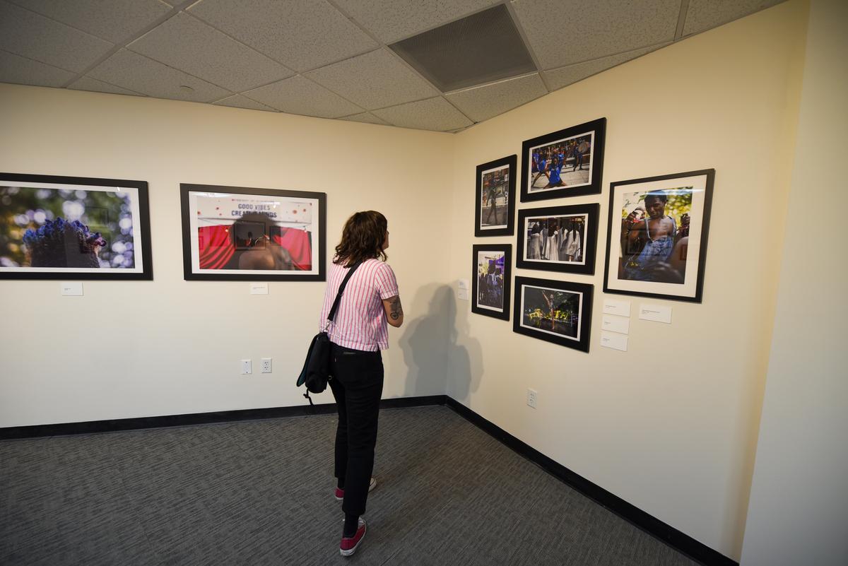 A person with shoulder-length brown hair, wearing a pink shirt and black pants, looks at a series of photographs framed on a cream-colored wall.