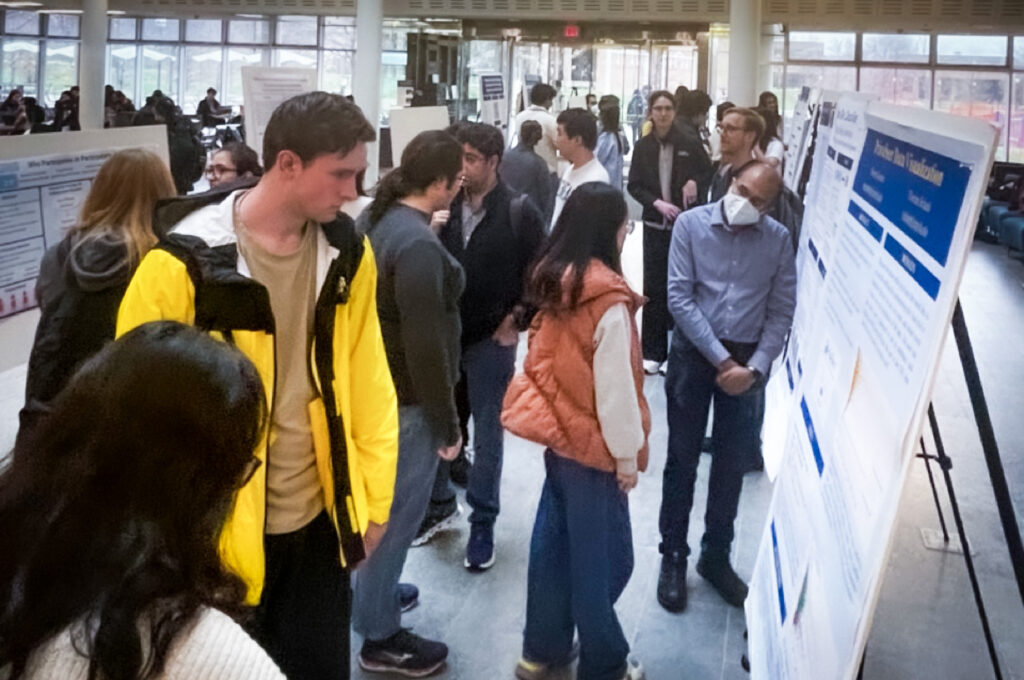 A crowd of around 20 people gathered around a row of research posters on stands, looking at posters and discussing with presenters.