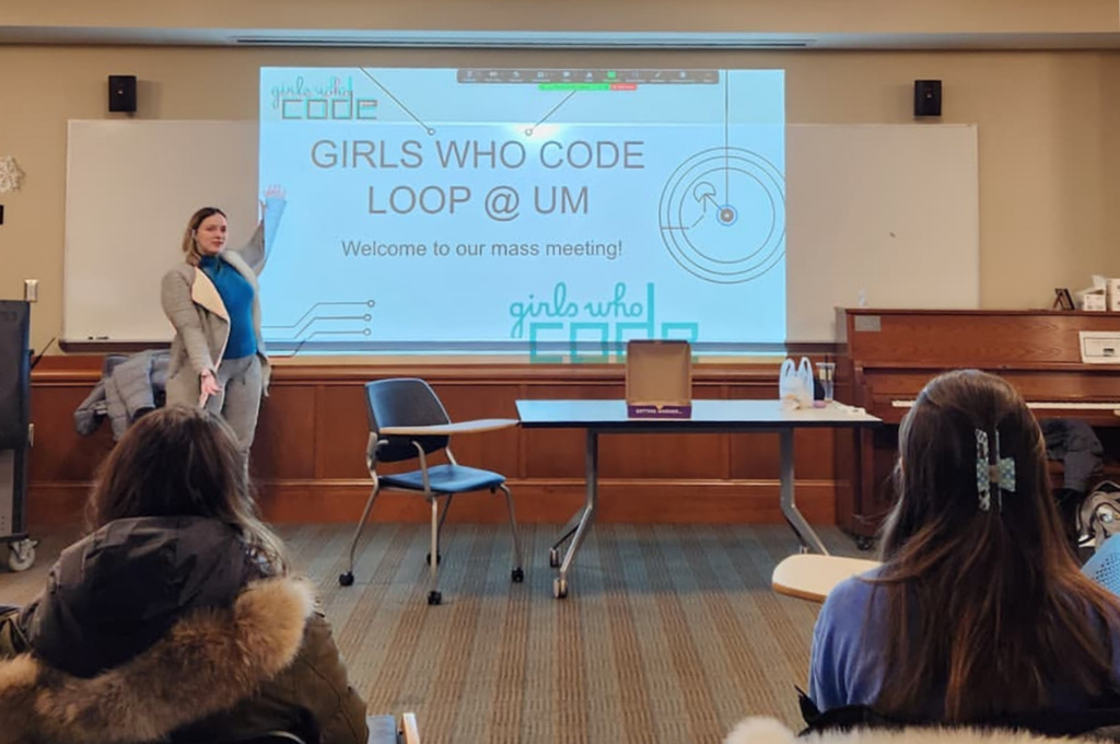 A woman presents a slideshow to a room of women students. On the slide, the text reads: "Girls Who Code Loop @ UM" and "Welcome to our mass meeting!"