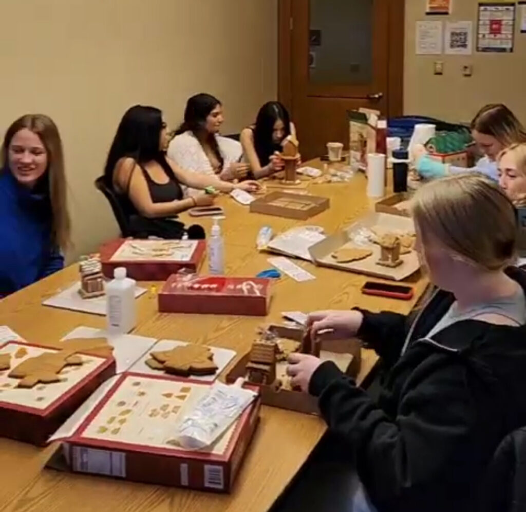 Several women sit at a long table chatting and building gingerbread houses