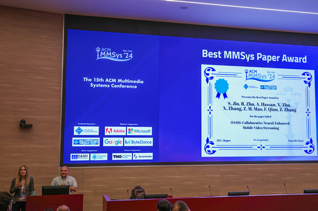 Two speakers stand at a podium. Behind them is a large projector screen announcing the Best MMSys Paper Award winner, with an image of a certificate with the OASIS team members and paper title listed.