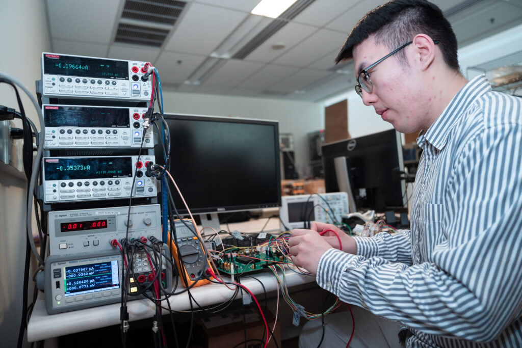 Qirui Zhang, wearing a blue and white vertically striped button up shirt and glasses, uses equipment in the lab. He sits next to a stack of electronic consoles displaying  voltage information.