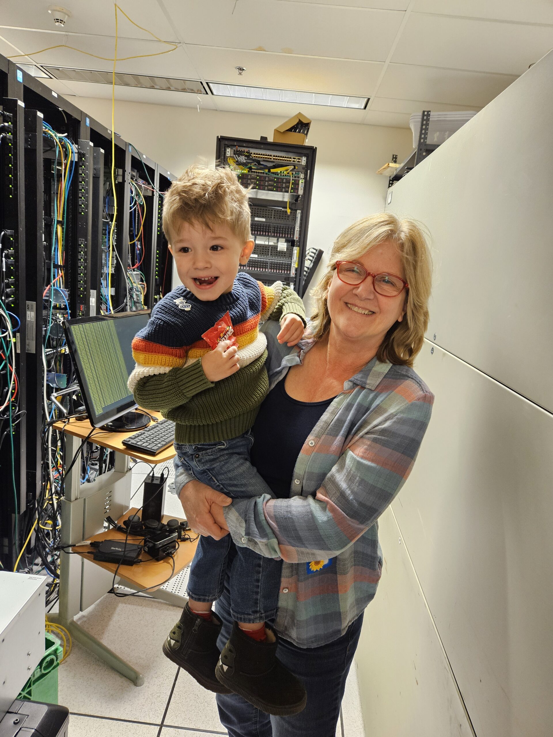 A blonde woman stands, holding a young child. They are both smiling at the camera. Behind them is a row of computer servers.