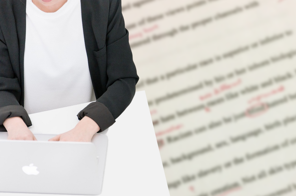 A photo of someone typing on a laptop - a front view of their torso (head is not included in image). In the background is a blurred image of a typed page with red proofreading marks.