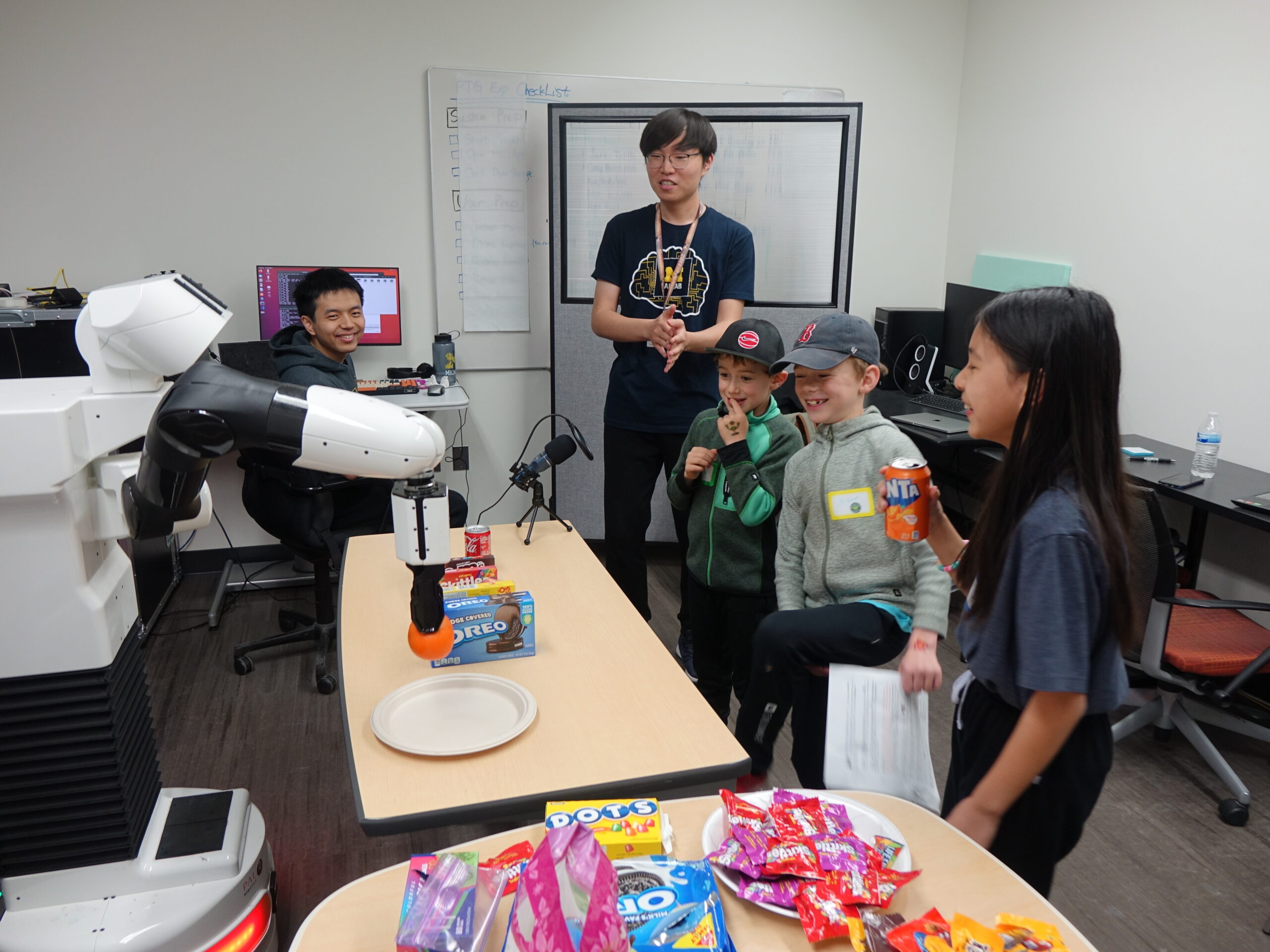 Three kids interact with a robot across a table as it selects and picks up items. A grad student stands nearby, supervising.