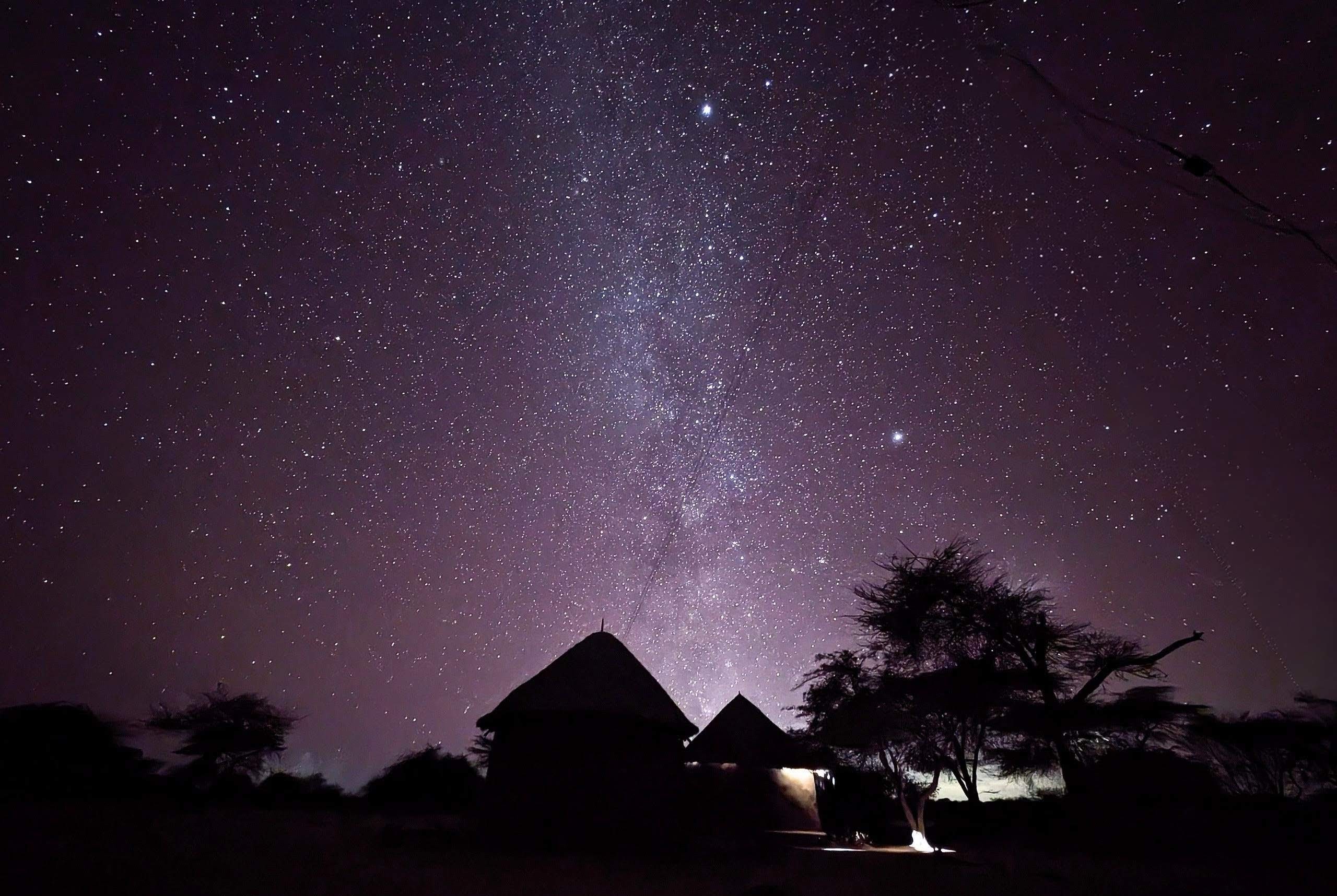 A photograph of the darkened sillouettes of trees and huts against the purple night sky, where the milky way is visible across the center of the photo.